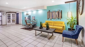 A spacious lobby with a yellow sofa, a blue armchair, and a coffee table. There is a bookshelf with books and decorative items, and a wall-mounted clock. - Days Inn by Wyndham Raleigh Glenwood-Crabtree (Raleigh)