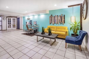 Un vestíbulo espacioso con un sofá amarillo, un sillón azul y una mesa de centro. Se aprecia una estantería con libros y objetos decorativos, así como un reloj de pared.