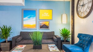 A living room with a sofa, a coffee table with magazines, two potted plants, and a blue armchair. - Days Inn by Wyndham Raleigh Glenwood-Crabtree (Raleigh)
