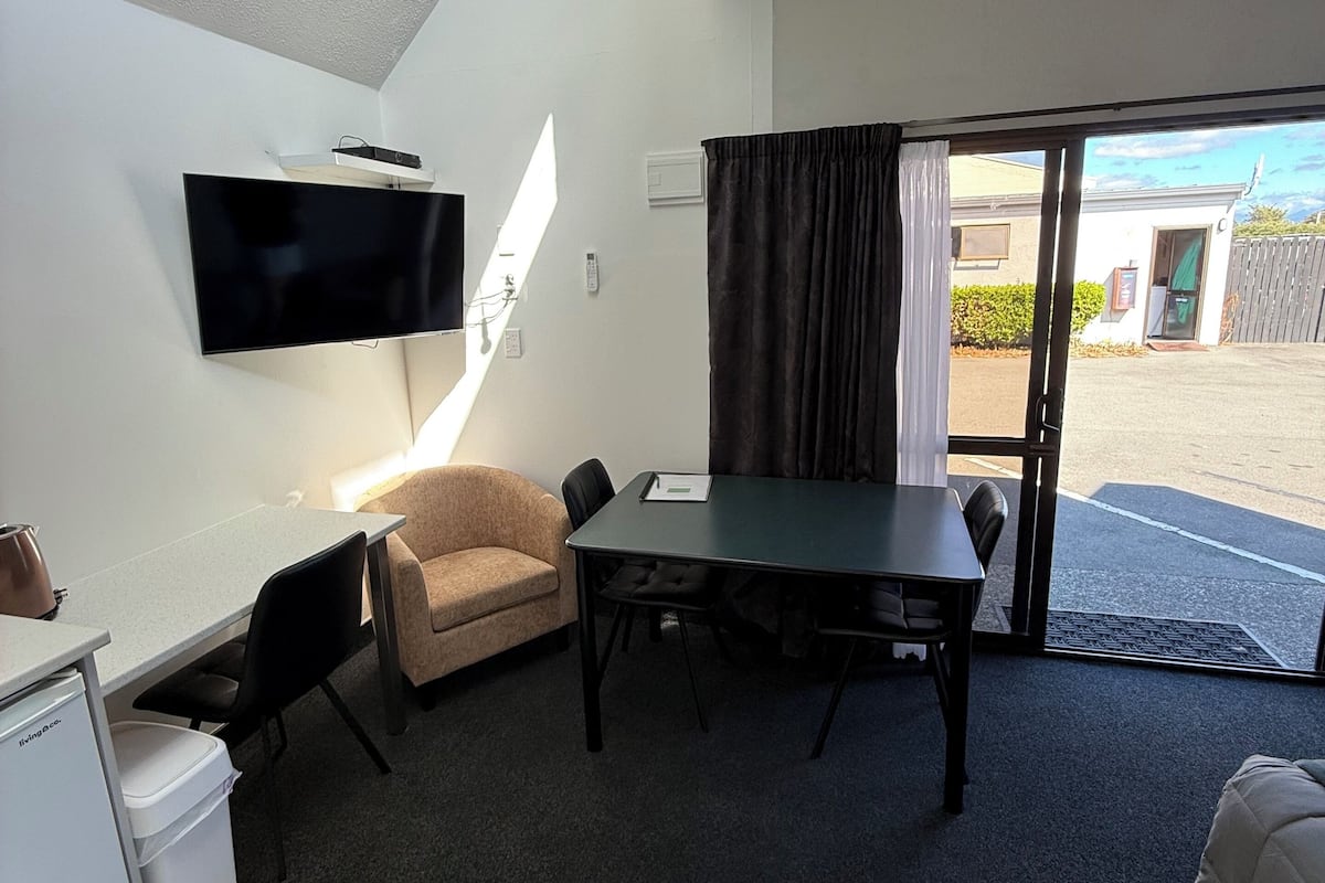 A room with a flat-screen TV mounted on the wall, a table with chairs, a sofa, and a sliding glass door leading to an outdoor area.