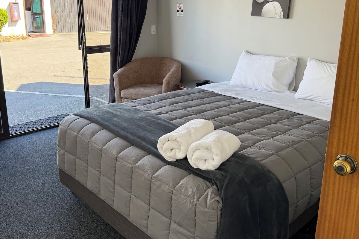 A hotel room with a bed, a chair, a painting, and an air conditioning unit.