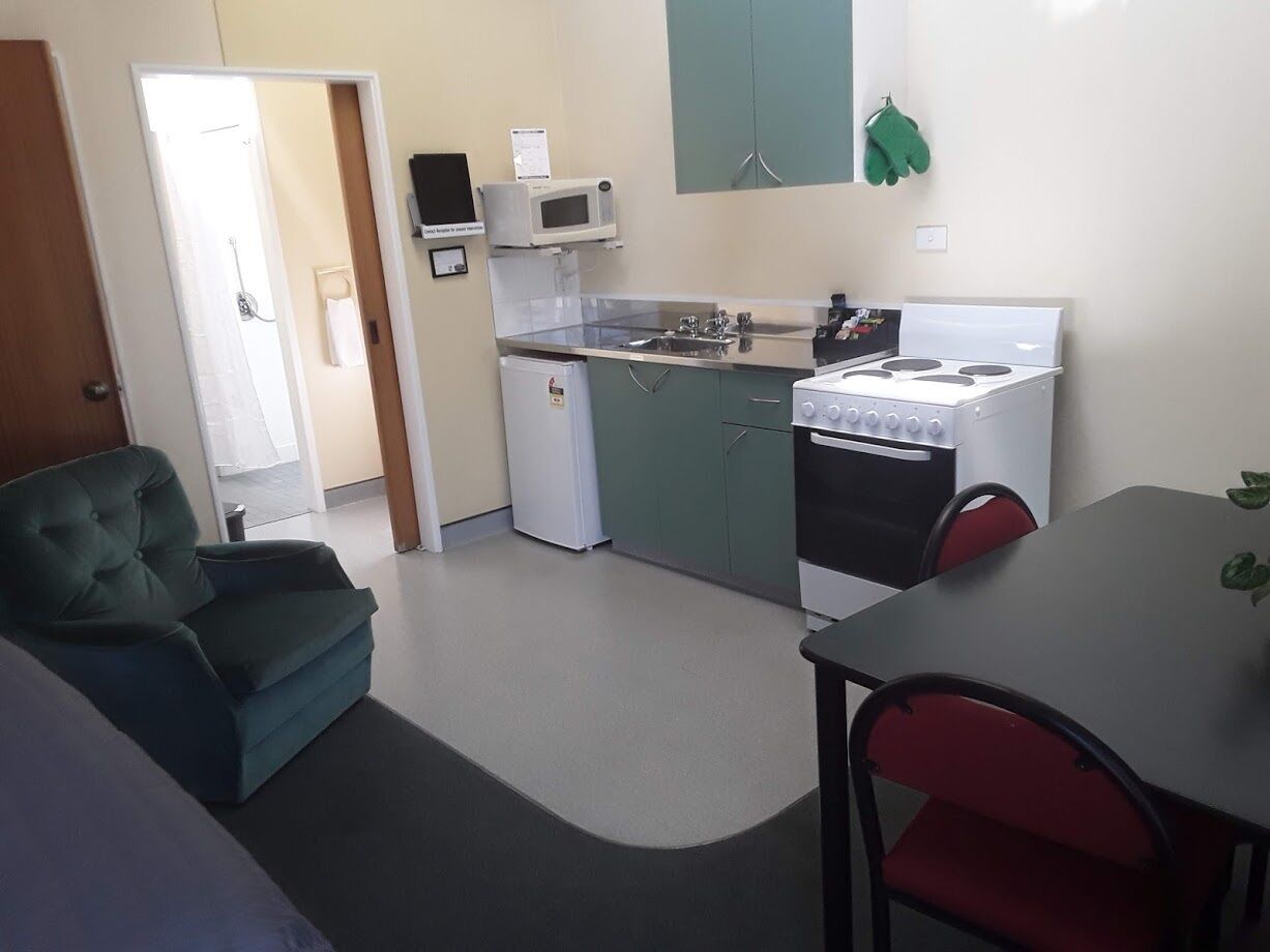 A compact kitchen with a microwave, stove, and refrigerator. There is a dining table with chairs and a green armchair.