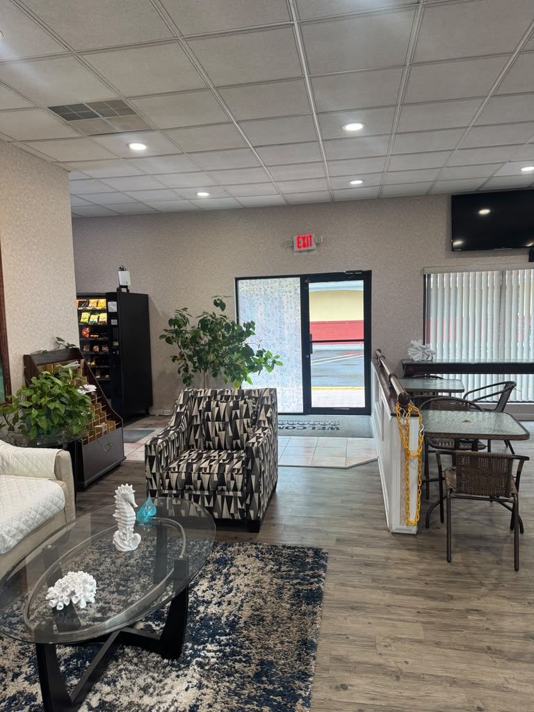 A lobby with a sofa, a patterned armchair, a glass coffee table, a dining area with a table and chairs, a bookshelf, and a large window with blinds.