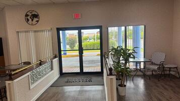 A hotel lobby with a reception desk, a world map on the wall, an exit sign, and a view of the street outside.