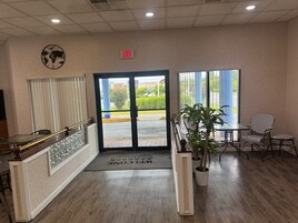 A hotel lobby with a reception desk, a world map on the wall, an exit sign, and a view of the street outside.