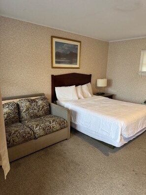A hotel room with a bed, a sofa, a nightstand, a lamp, and a framed picture on the wall.