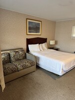 A hotel room with a bed, a sofa, a nightstand, a lamp, and a framed picture on the wall.