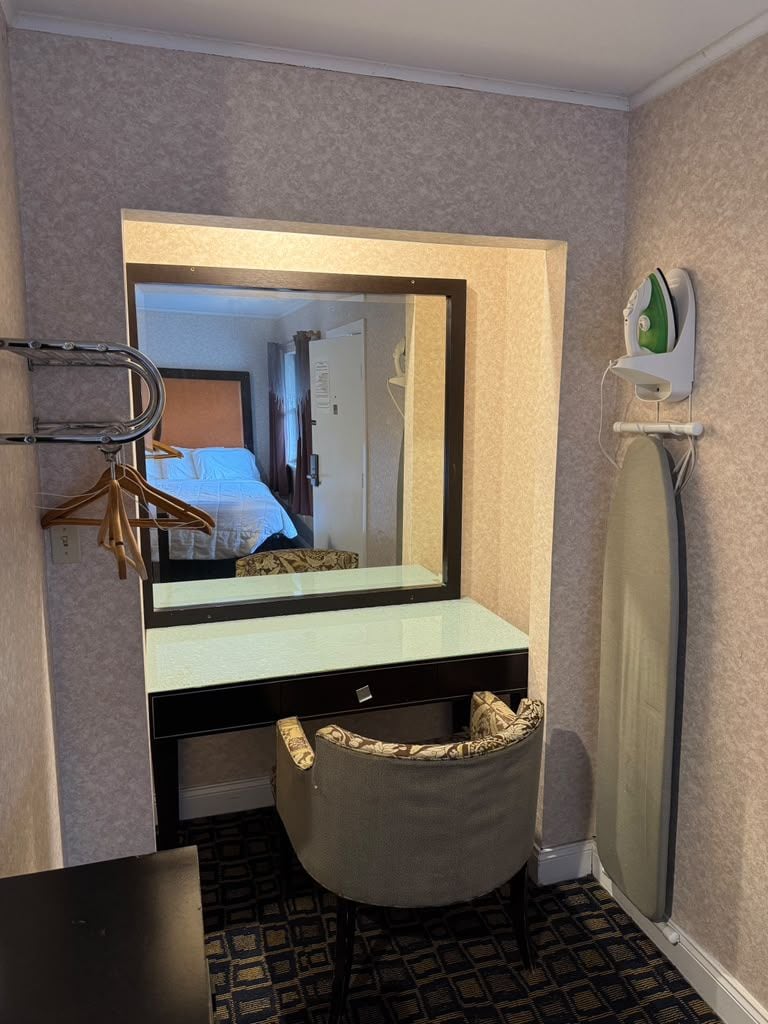 Een kleine hotelkamer met een spiegel, een klein kaptafeltje, een kledingrek, een koffer en een wandijzer.