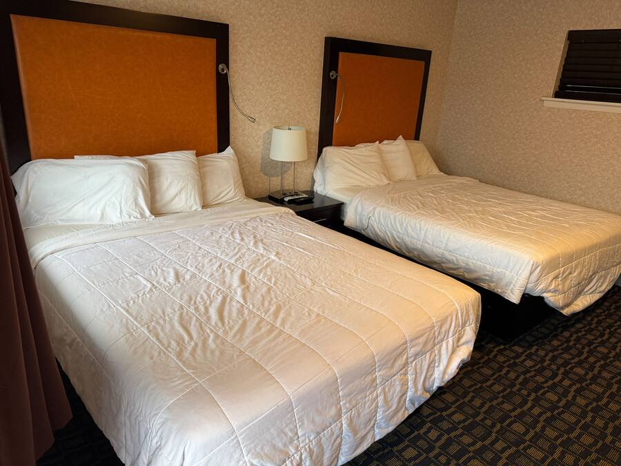 A hotel room with two beds, a nightstand with a lamp, and a window with blinds.