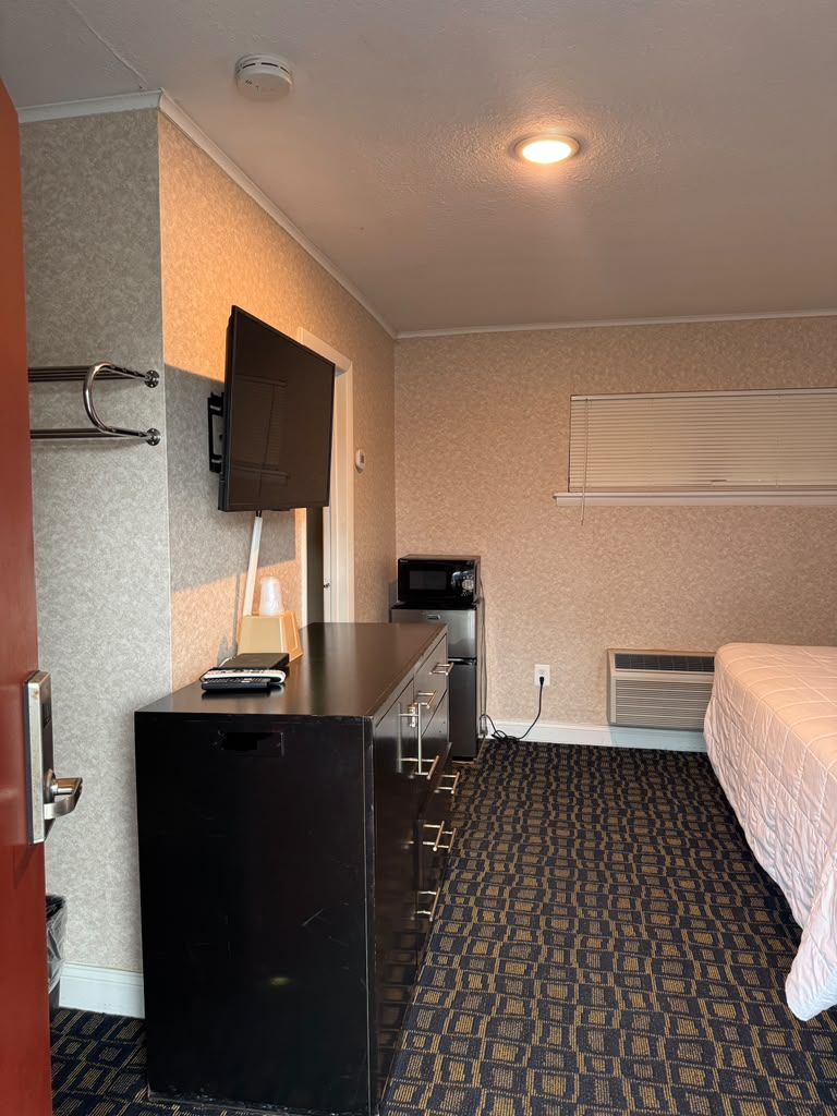 A hotel room with a bed, a desk with drawers, a television mounted on the wall, and a window with blinds.