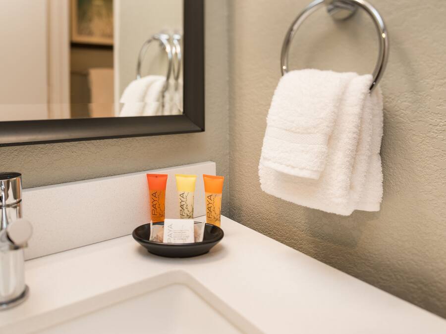A bathroom sink with a mirror, a towel hanging on a metal ring, and three tubes of lotion.