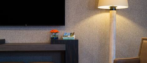 A modern living room with a flat-screen TV mounted on the wall, a tall floor lamp, and a dark wooden coffee table with books and a vase of flowers.