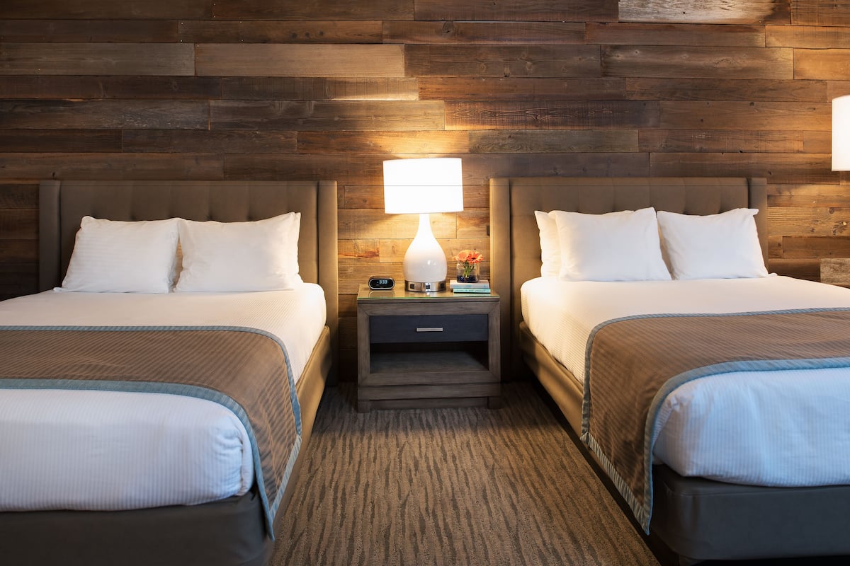 A hotel room with two beds, a nightstand with a lamp, and a wooden wall.