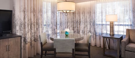 Habitación de hotel con una mesa de comedor redonda, dos sillas, un televisor sobre un mueble bajo y una lámpara en una mesita.