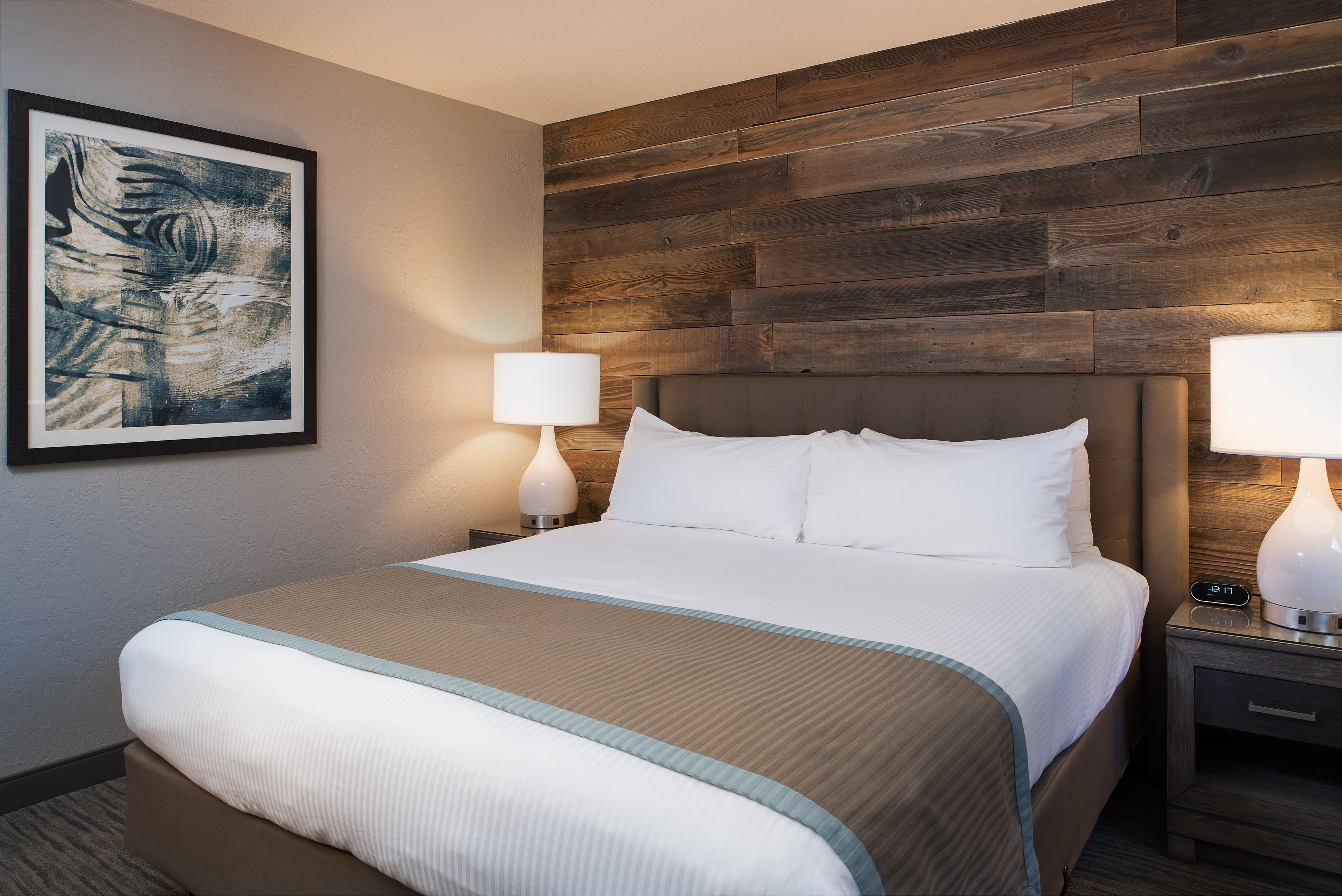 Ein Hotelzimmer mit einem großen Bett, zwei Nachttischen mit Lampen, einem hölzernen Kopfteil und einem gerahmten, abstrakten Gemälde an der Wand.