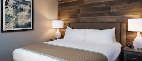 Une chambre d’hôtel avec un grand lit, deux tables de chevet avec des lampes, une tête de lit en bois et un tableau abstrait encadré accroché au mur.