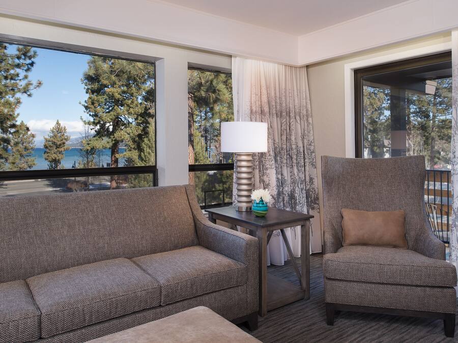 Habitación de hotel con sofá, sillón y una mesa con lámpara. Se ven árboles y montañas a través de las ventanas.