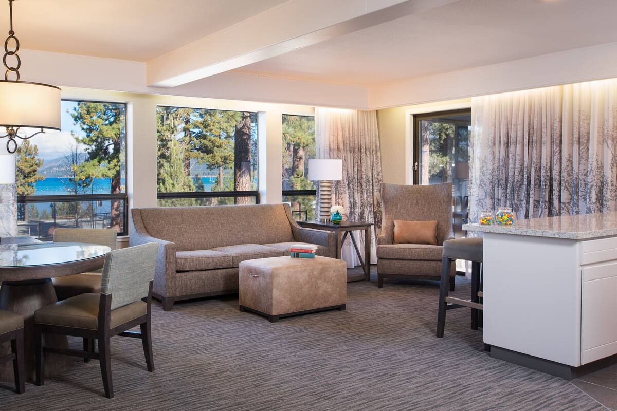 A modern hotel room with a large window offering a scenic view, a cozy seating area with a sofa and ottoman, a small table, and a bar area.