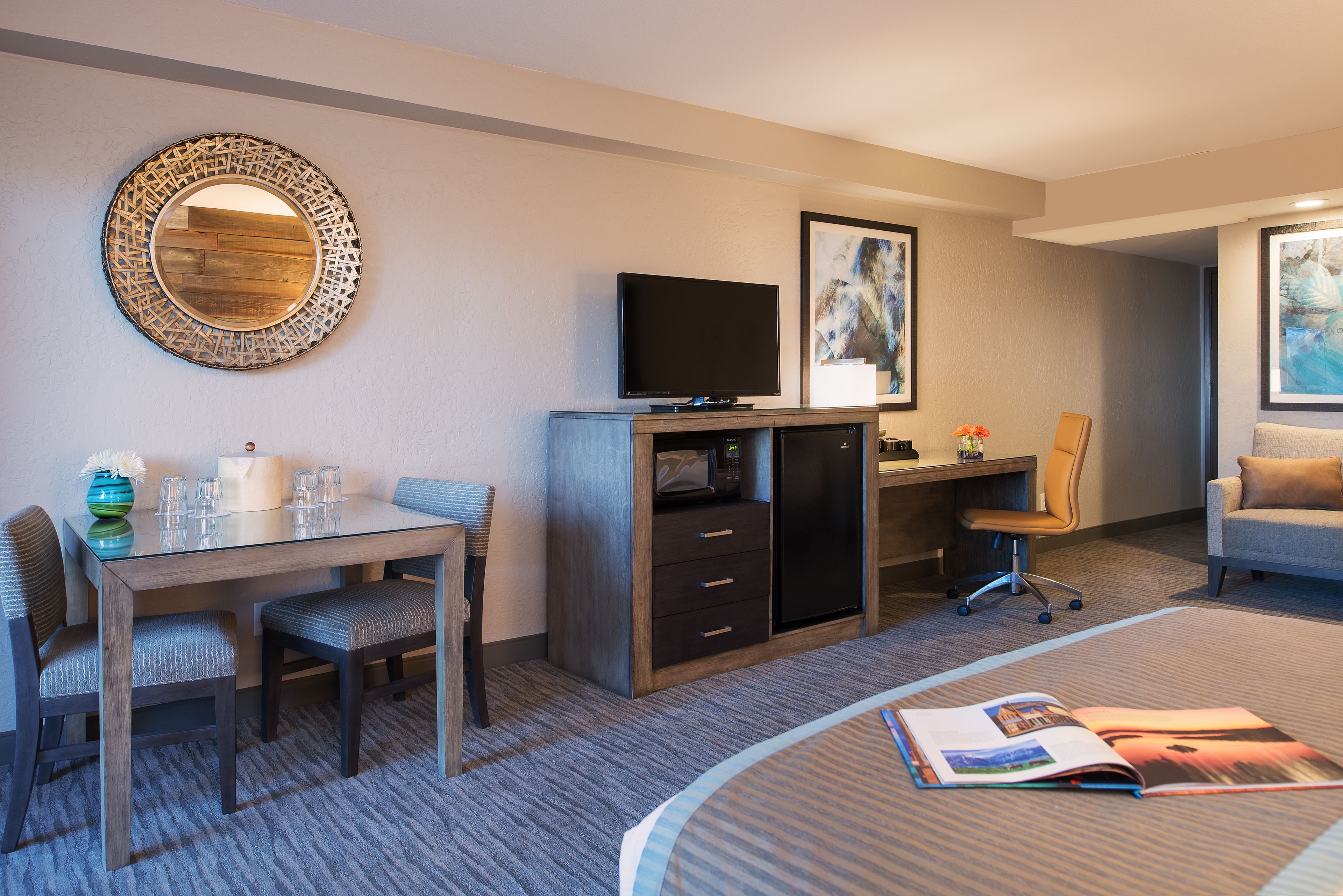 A hotel room with a dining table, chairs, a flat-screen TV, a microwave, a desk with a computer, a chair, a sofa, and a magazine on the floor.