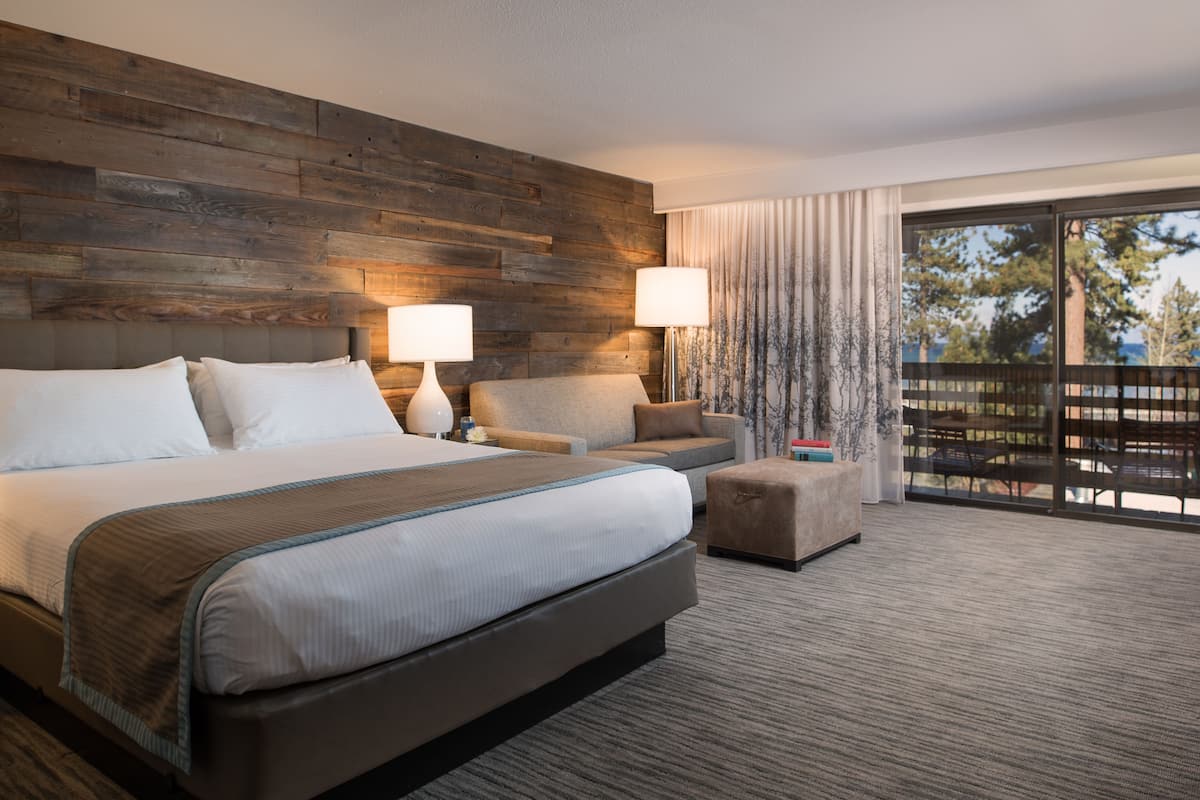 A modern hotel room with a large bed, two bedside lamps, a sofa, and a balcony with a view of trees.