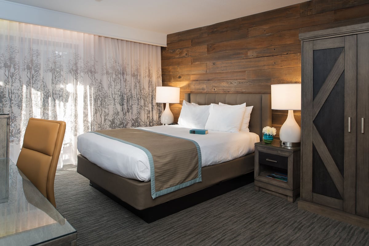 A hotel room with a large bed, two bedside tables with lamps, a wooden headboard, and a view of trees through sheer curtains.