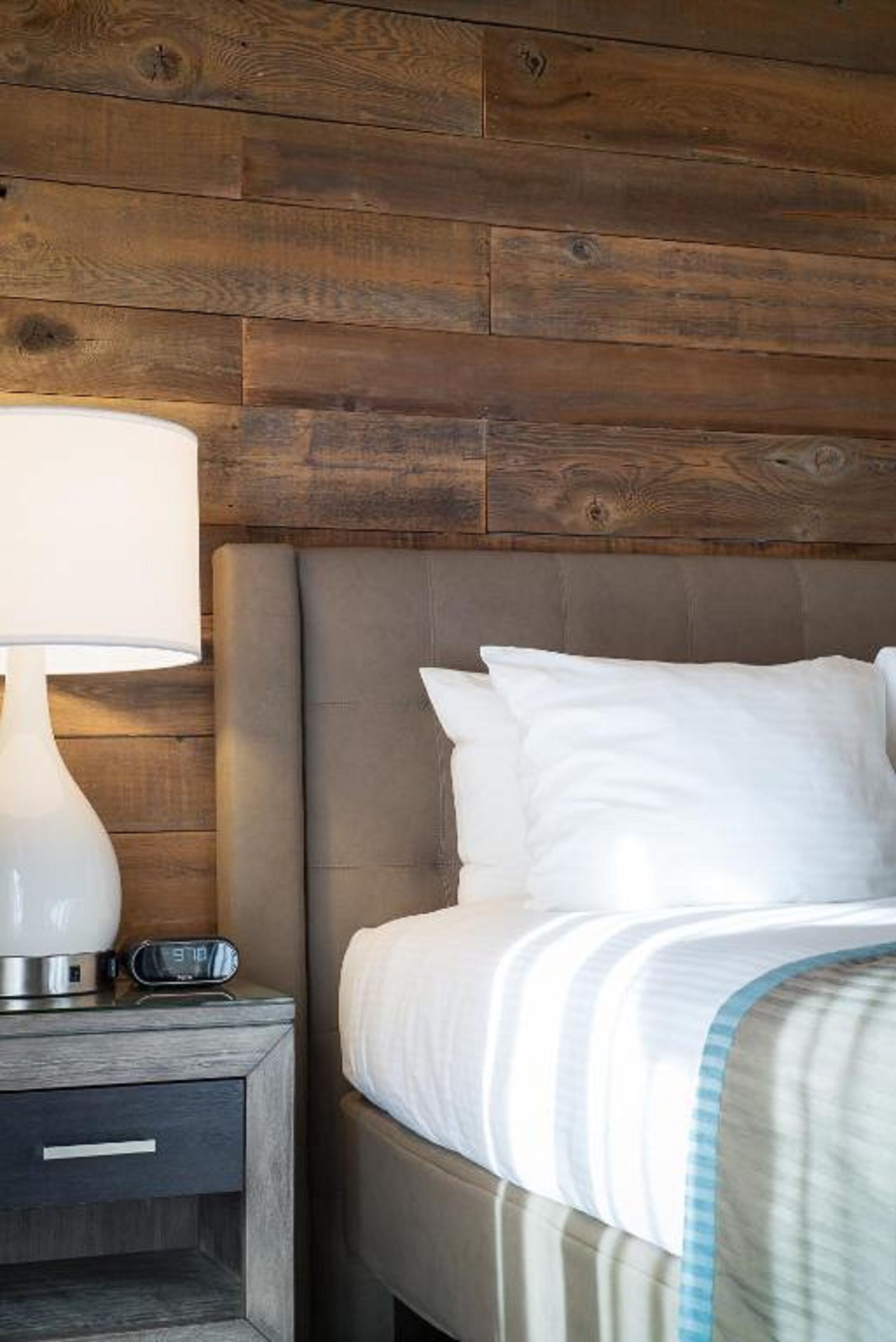 A neatly made bed with white bedding and a striped blanket, a bedside lamp, and a digital alarm clock on a wooden nightstand.