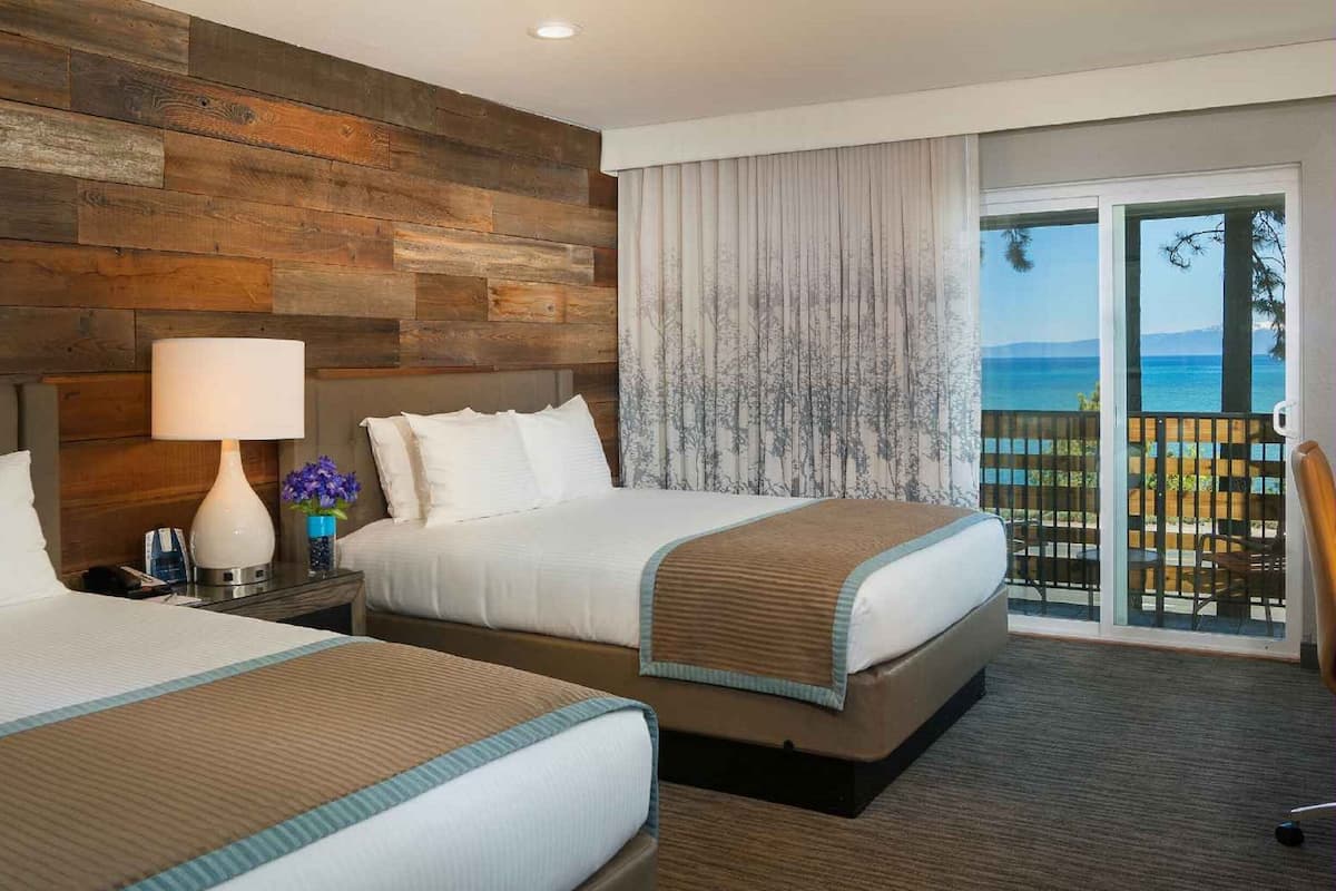 A hotel room with two beds, a wooden wall, a balcony with a view, and a nightstand with a lamp and vase.