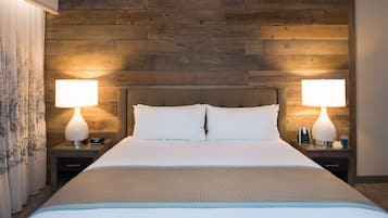 Une chambre d’hôtel avec une tête de lit en bois, deux lampes de chevet, un lit avec du linge de lit blanc et une table de chevet avec une horloge et une bouteille.