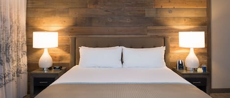 Una habitación de hotel con cabecera de madera, dos lámparas de noche, una cama con ropa blanca y una mesita de noche con un reloj y una botella.