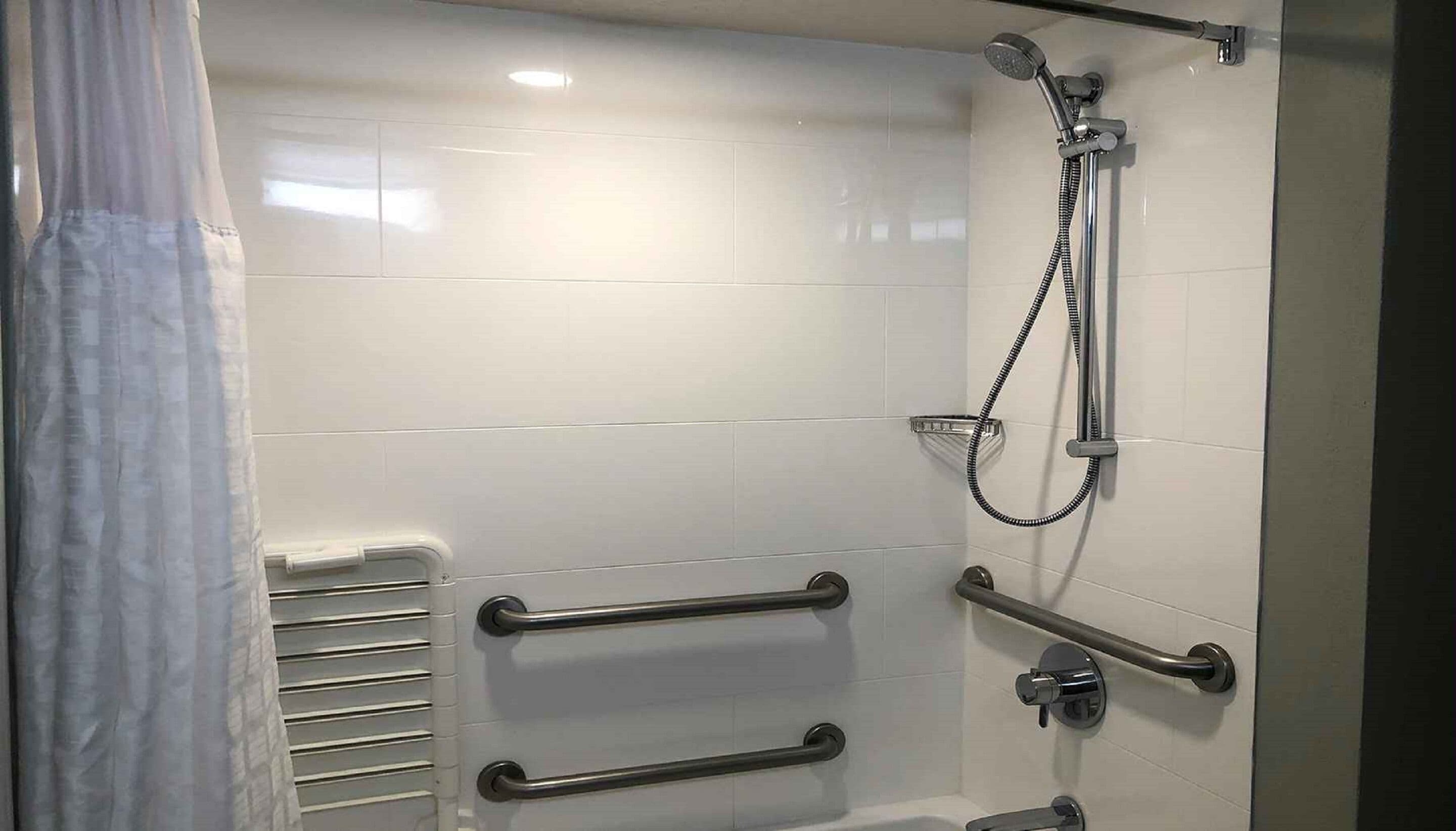 A modern bathroom with white tiles, a shower with a handheld hose, and two grab bars for support.