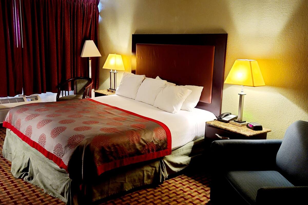 A hotel room with a large bed, two bedside lamps, a chair, and a desk with a phone and a clock.