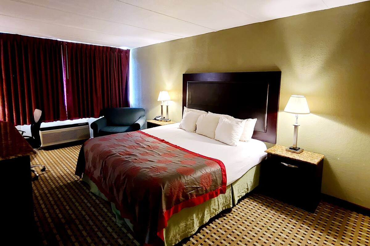 A hotel room with a bed, two bedside lamps, a chair, a desk, and a heater.