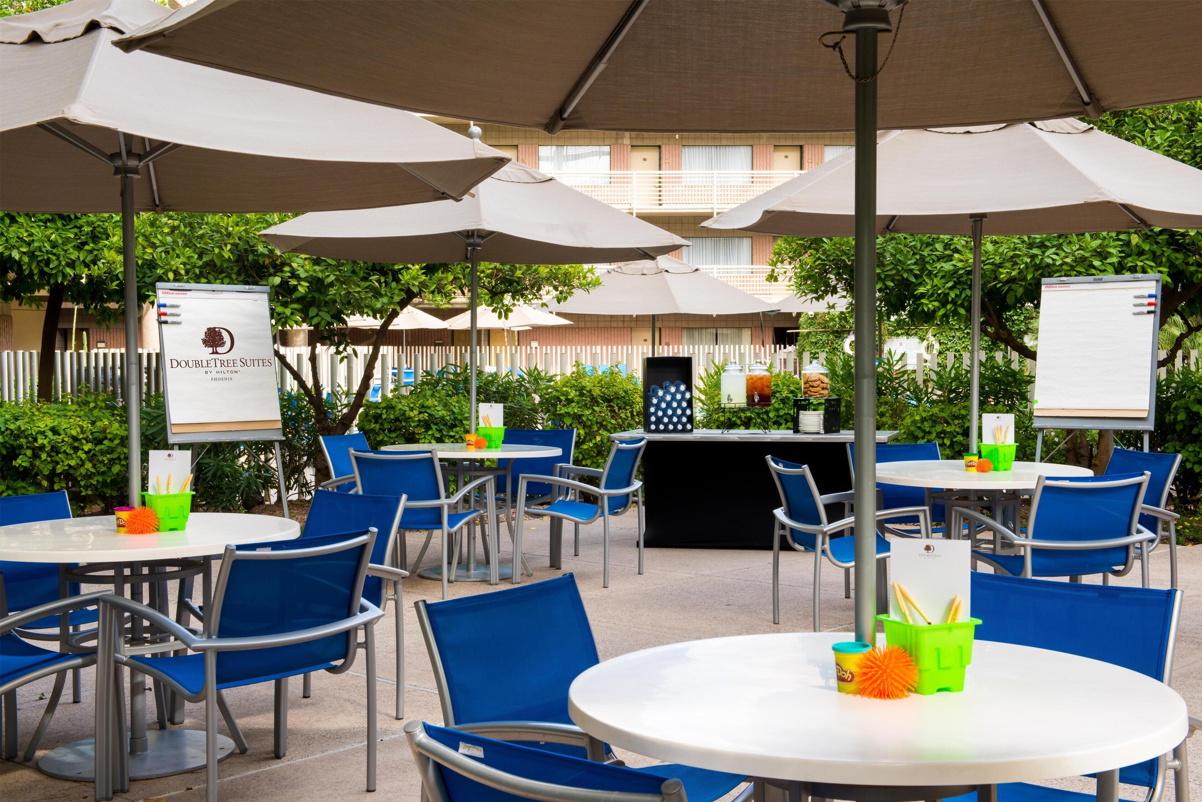 An outdoor dining area with round tables and blue chairs, shaded by large umbrellas, and a signboard for DoubleTree Suites.