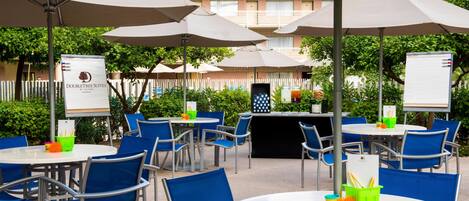 An outdoor dining area with round tables and blue chairs, shaded by large umbrellas, and a signboard for DoubleTree Suites.