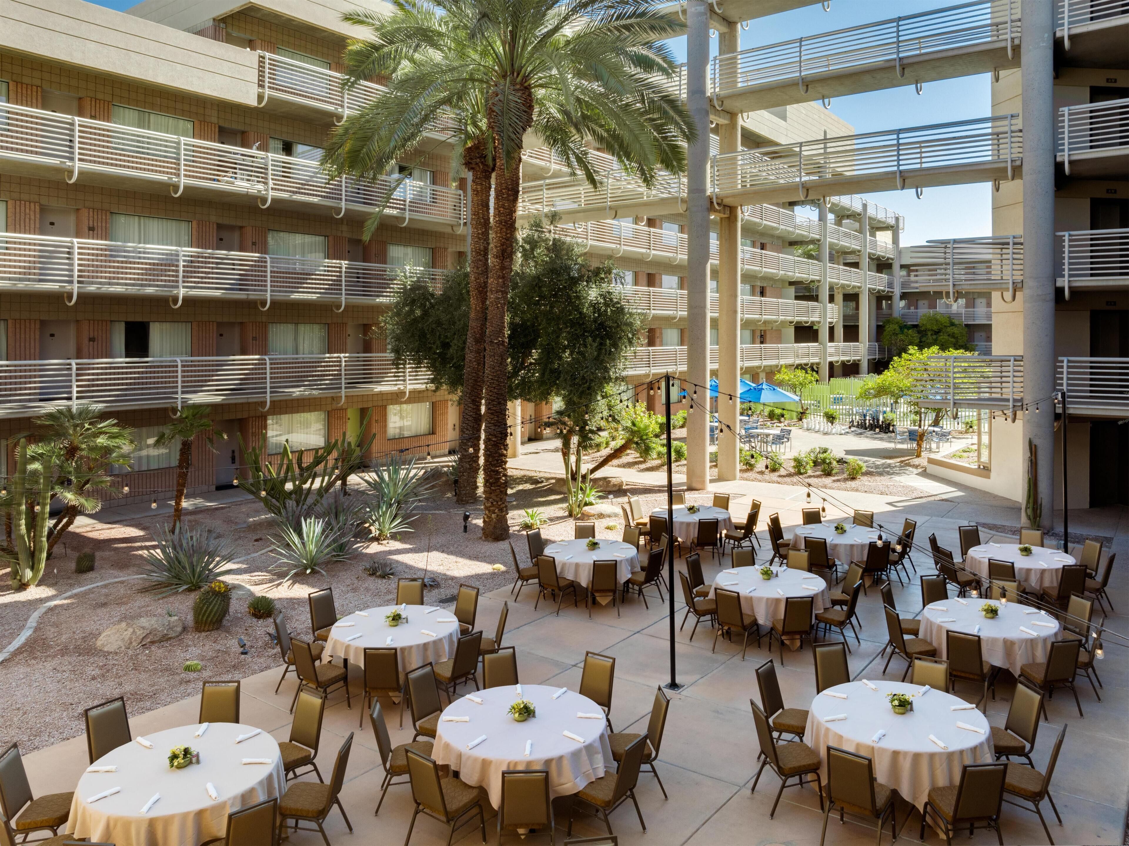 A hotel courtyard with outdoor dining tables, palm trees, and a multi-story building with balconies.