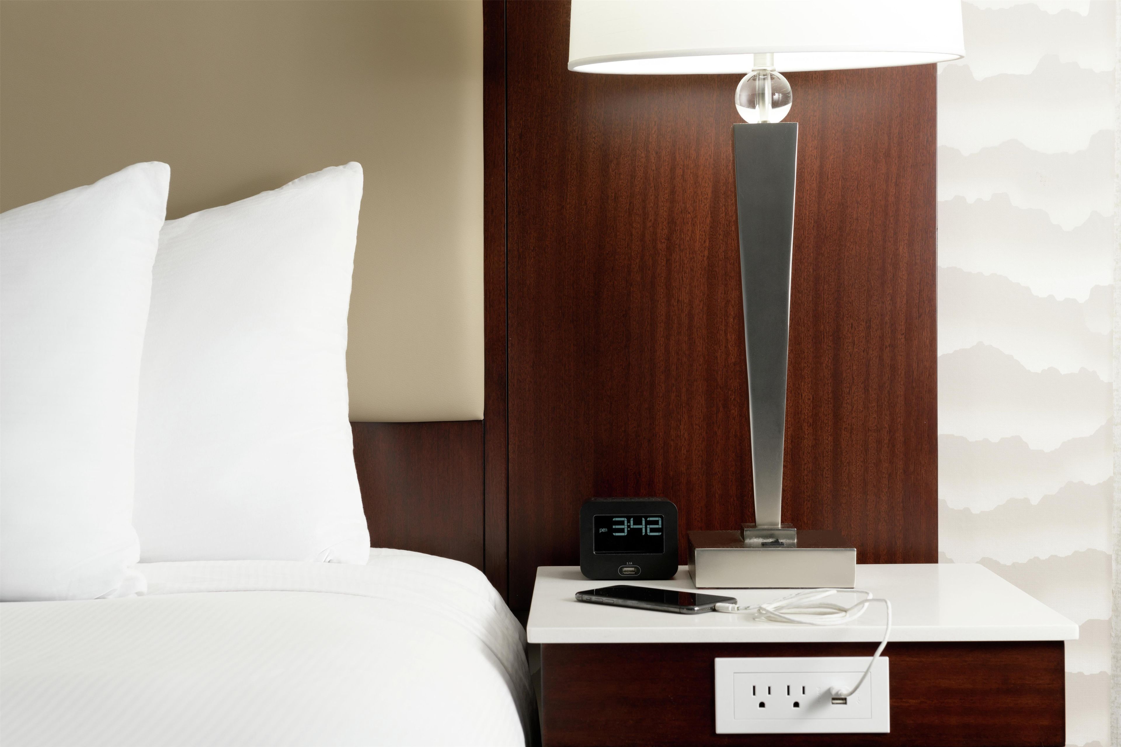 Una habitación de hotel con una cama, una mesita de noche con un reloj digital, una lámpara y un enchufe.