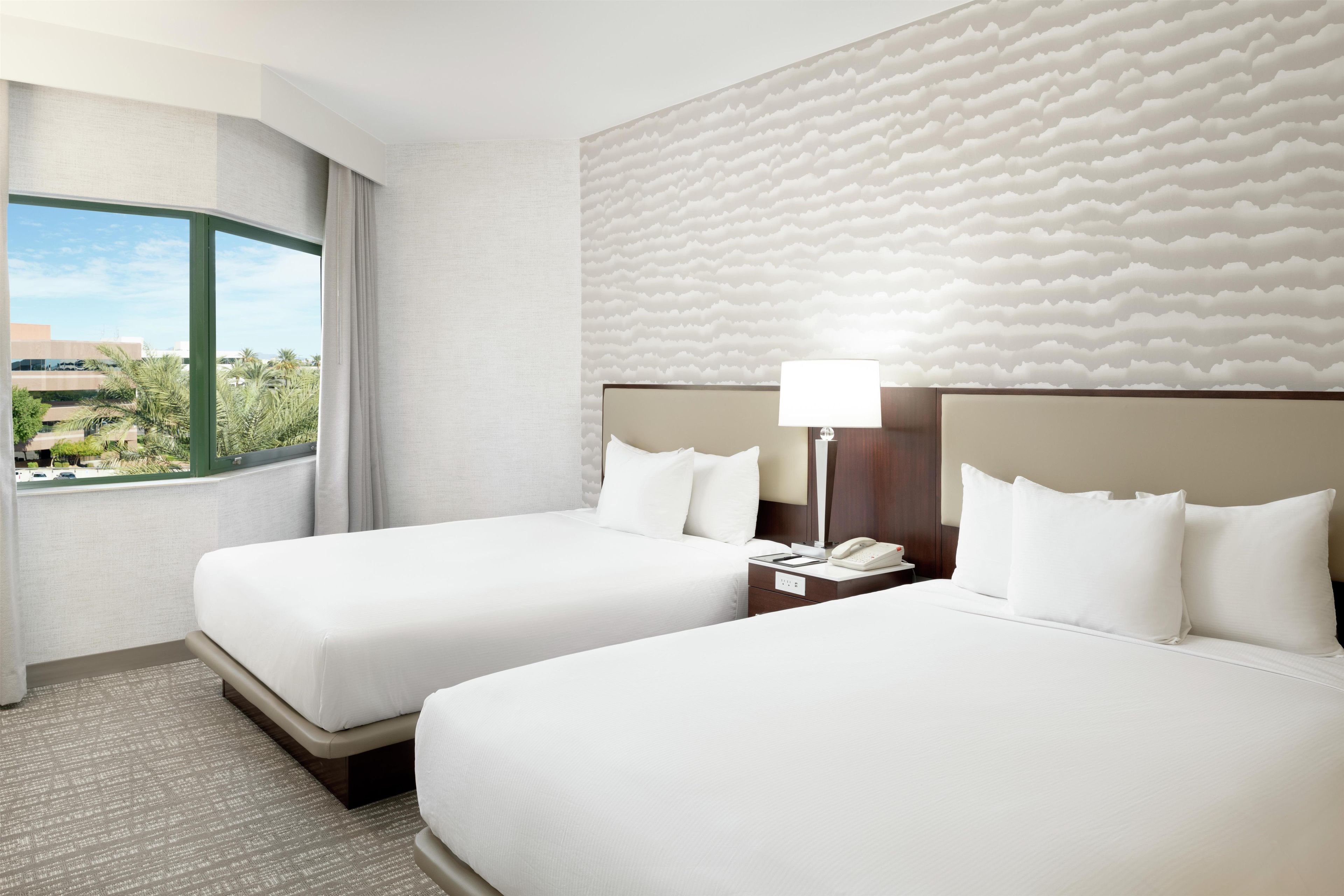 A hotel room with two beds, a view of the outdoors, a textured wall, and a nightstand with a lamp.