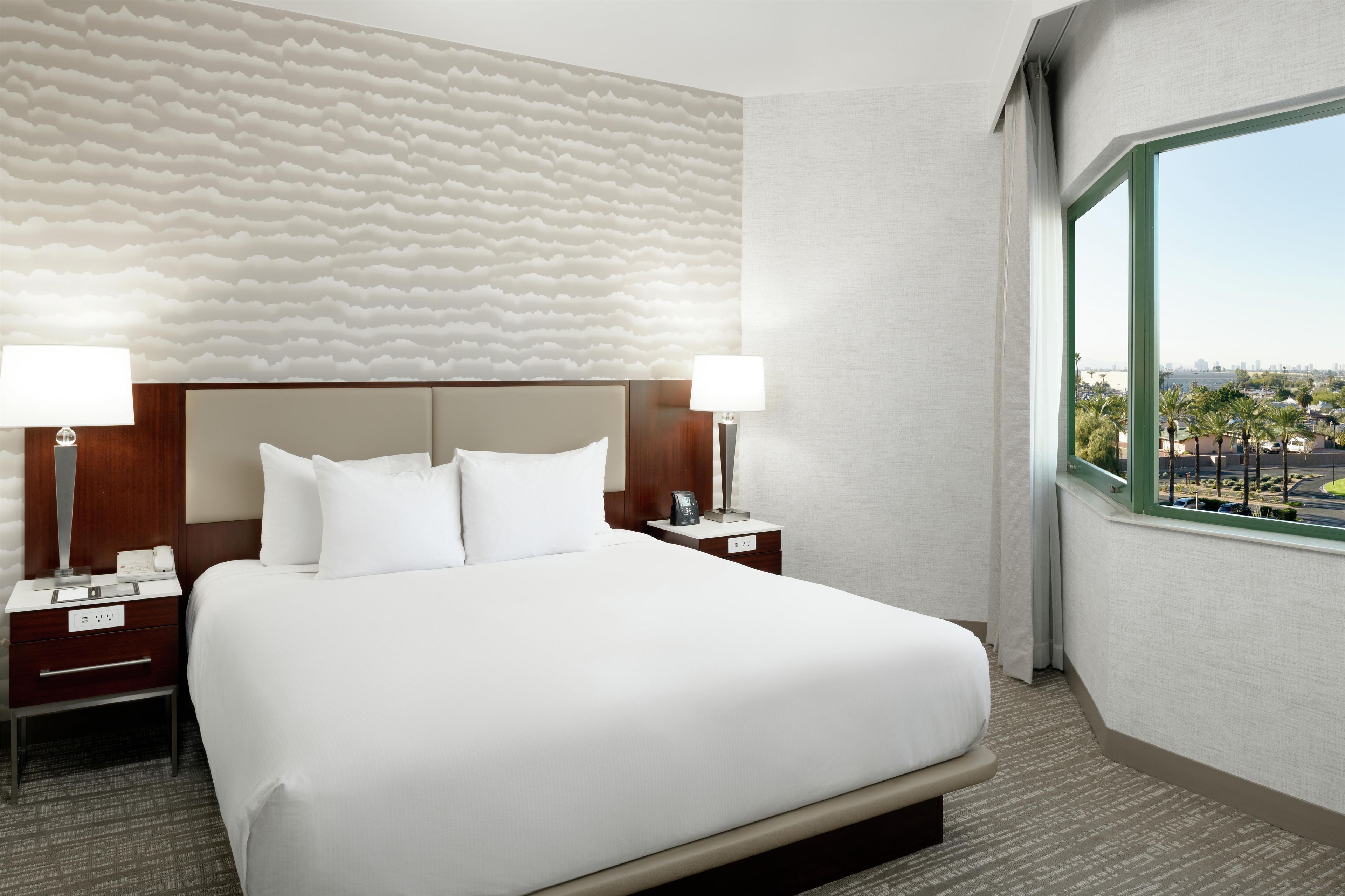 A hotel room with a large bed, two bedside tables, a headboard, and a view of palm trees and a cityscape through a window.