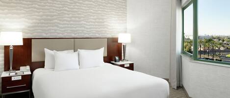 A hotel room with a large bed, two bedside tables, a headboard, and a view of palm trees and a cityscape through a window.