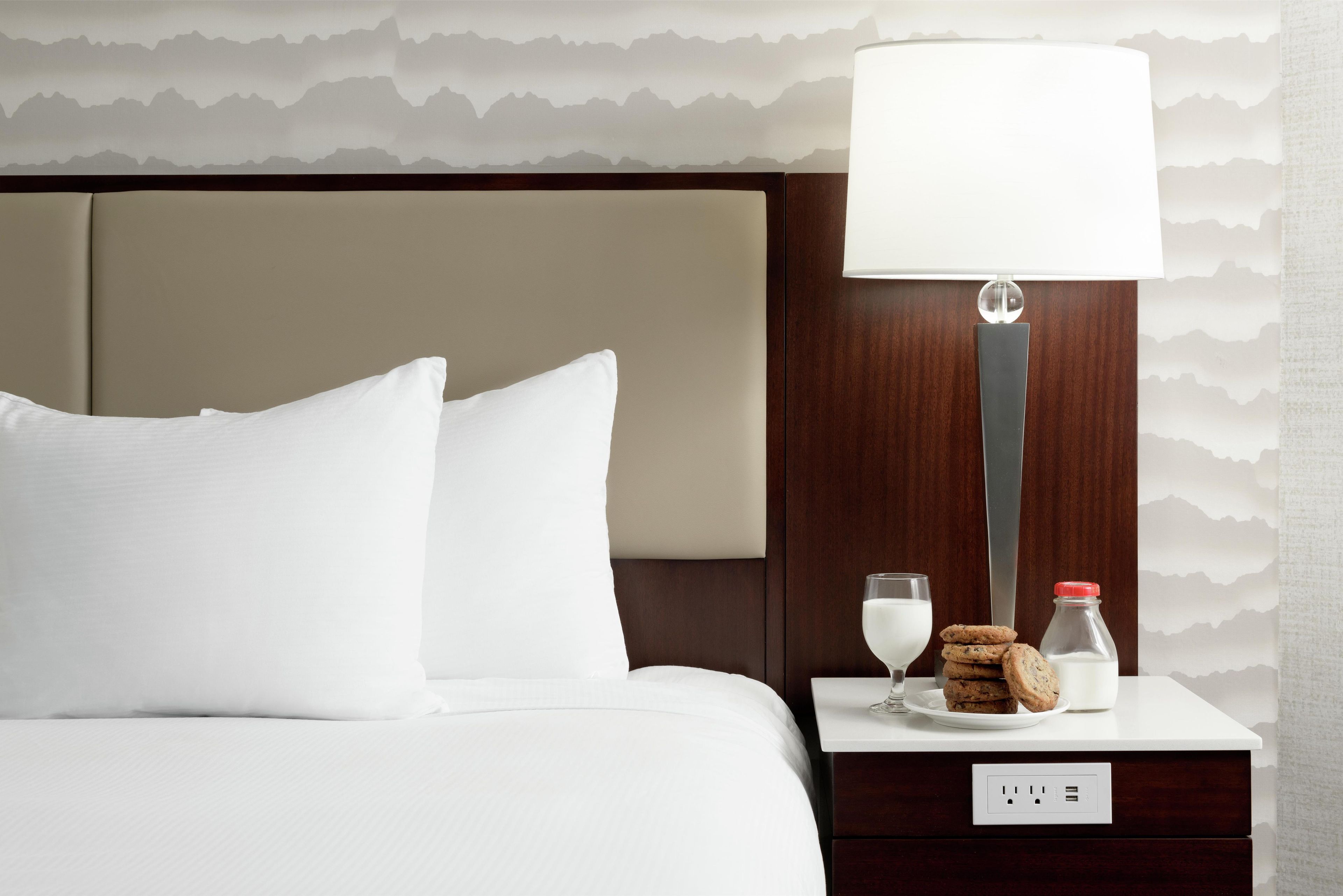 A hotel room with a bed, a nightstand with a lamp, a glass of milk, cookies, and a milk bottle.