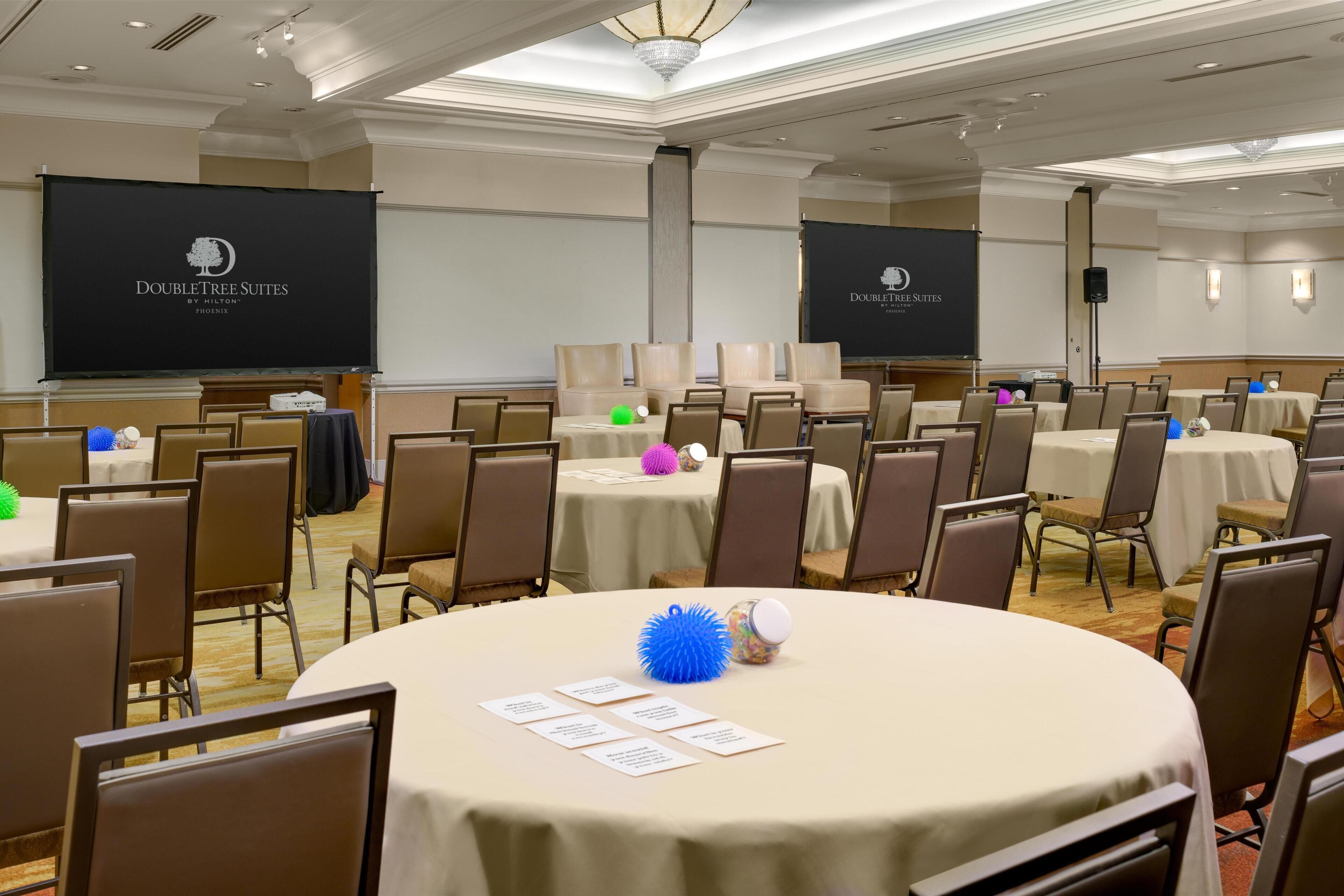 Sala de conferência com mesas redondas, cadeiras e telas de projeção exibindo o logotipo da DoubleTree Suites.