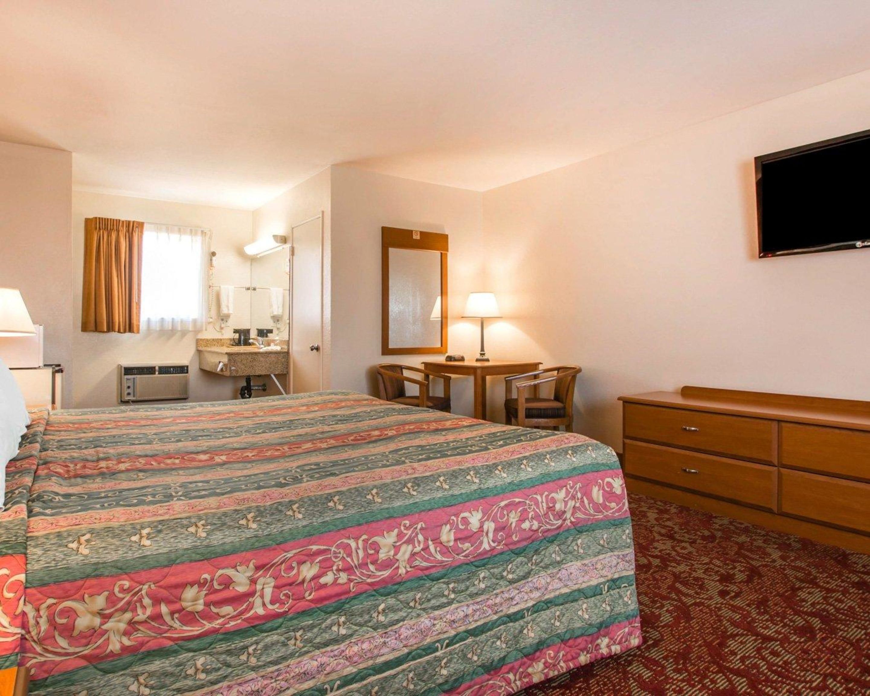 A hotel room with a bed, a desk, a chair, a dresser, a television, and a bathroom with a mirror and a window.