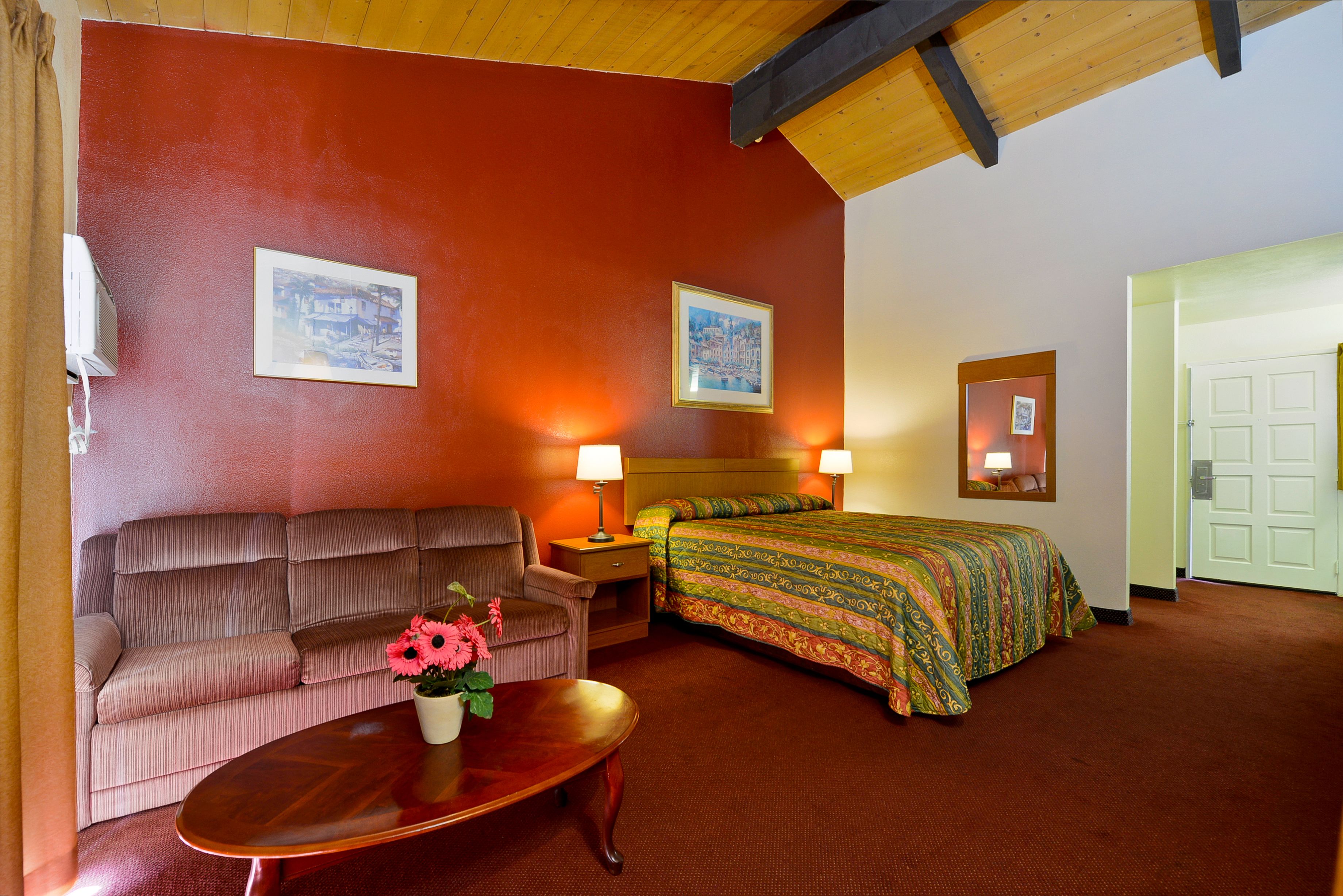 A hotel room with a sofa, a bed with a colorful bedspread, a wooden coffee table with a flower vase, and two wall-mounted lamps.