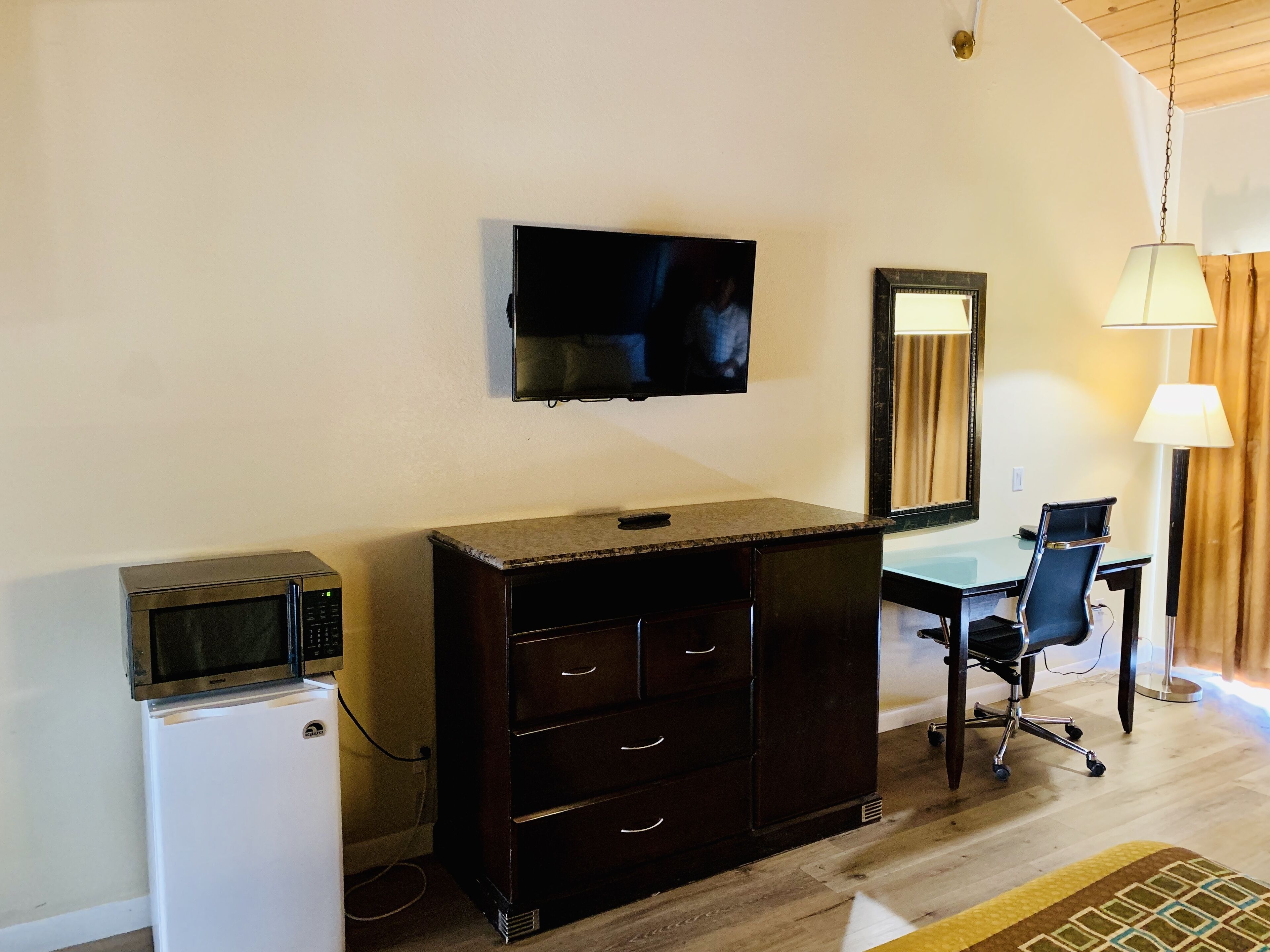 Una habitación con televisión montada en la pared, un escritorio con silla, un refrigerador pequeño y un aparador de madera oscura.