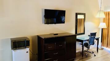 Una habitación con televisión montada en la pared, un escritorio con silla, un refrigerador pequeño y un aparador de madera oscura.