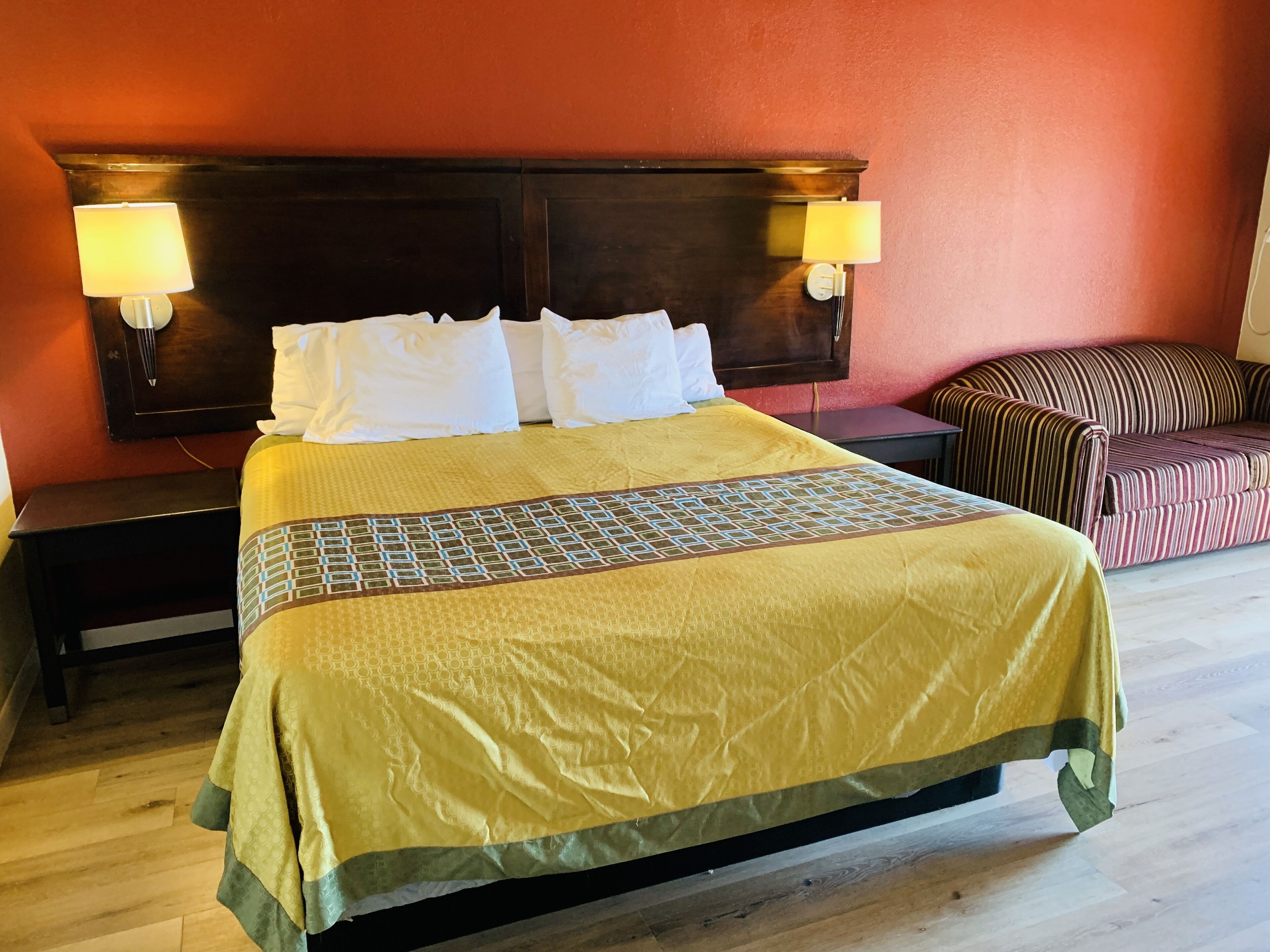 A hotel room with a large bed, two bedside lamps, a wooden headboard, a striped sofa, and a wooden floor.