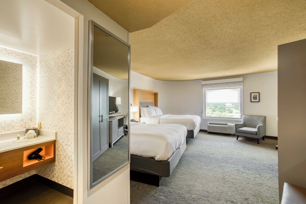 A hotel room with two beds, a bathroom with a vanity, a window with a view, and a chair.