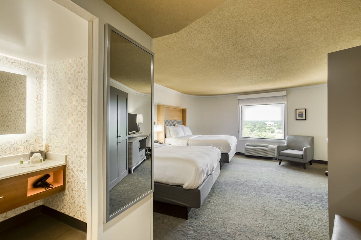 A hotel room with two beds, a bathroom with a vanity, a window with a view, and a chair.
