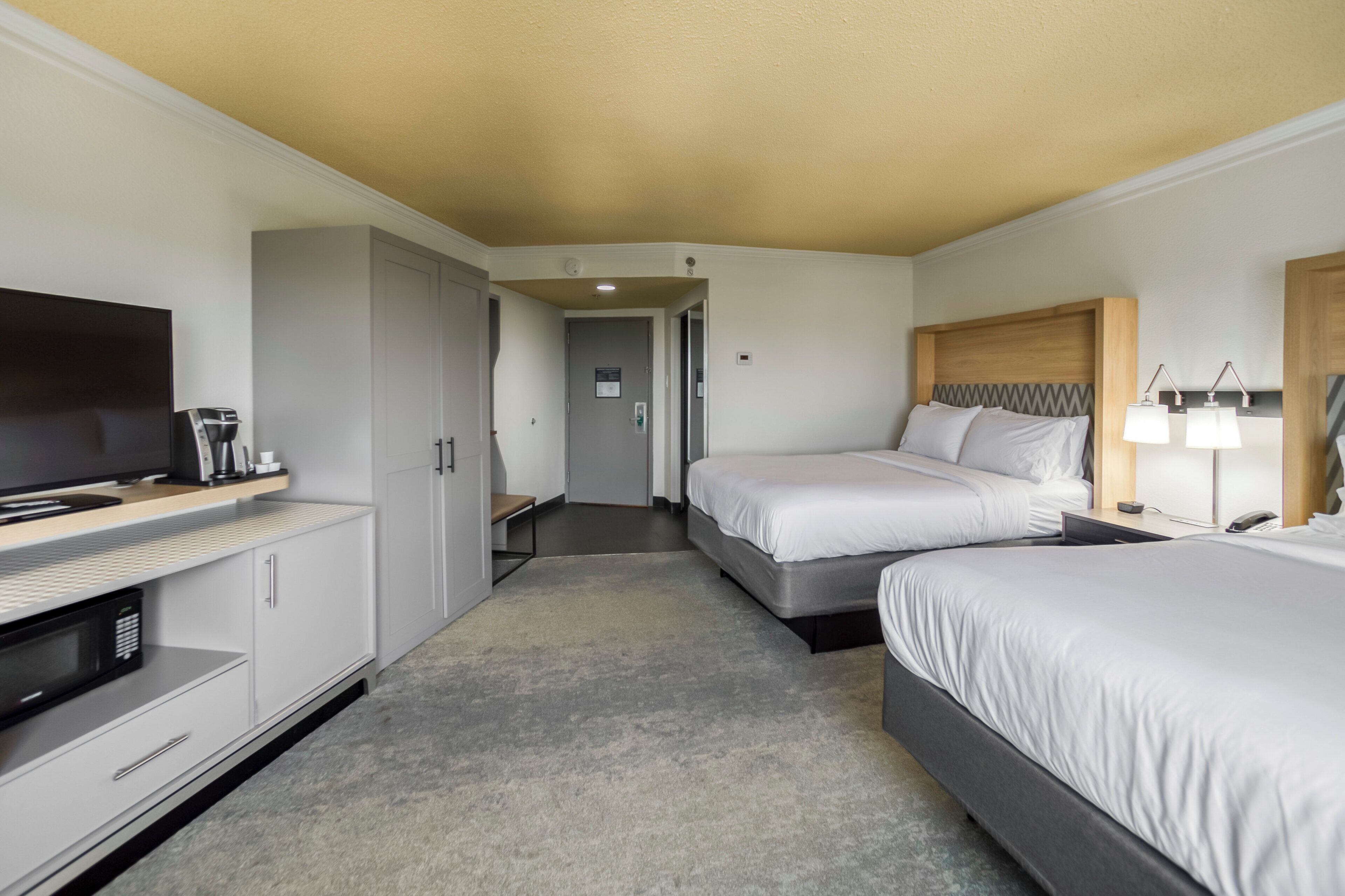 Habitación de hotel con cama, mesita de noche, lámpara, televisor y escritorio con computadora y cafetera.
