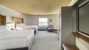 Ein Hotelzimmer mit zwei Betten, einem Schreibtisch, einem Sessel, einem Fernseher und einem Fenster mit Blick auf Grünflächen.
